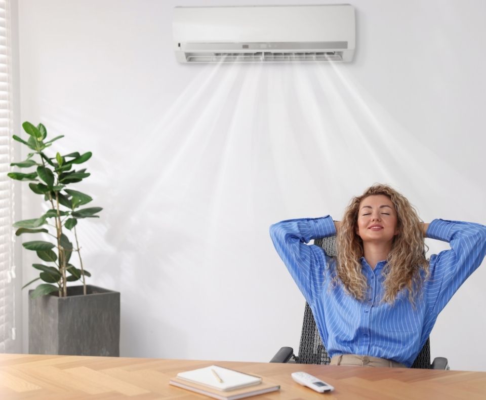 Woman relaxing in an office chair with hands behind head, enjoying cool air from a wall-mounted AC unit above her.