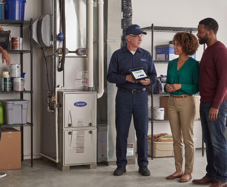 HVAC technician in navy uniform shows a tablet to a couple in a garage beside a Carrier furnace.