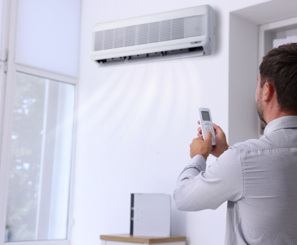 Man holding a remote and aiming it at a wall-mounted air conditioner in a bright room.]​;