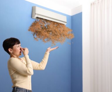 Woman in a beige sweater blows air from a wall-mounted air conditioner, dust or leaves puffing out toward the blue wall.