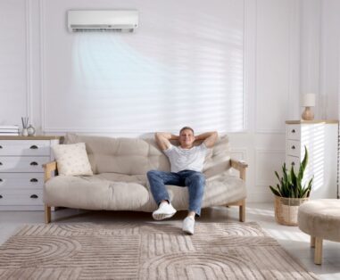 Man relaxing on a beige sofa in a bright living room while the wall AC blows cool air above him.