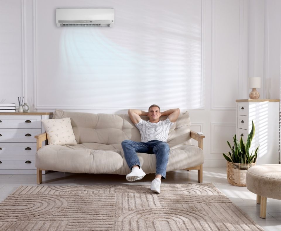 Man relaxing on a beige sofa in a bright living room while the wall AC blows cool air above him.
