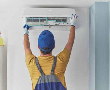 HVAC technician installing a wall-mounted air conditioner, wearing gloves and a blue cap.