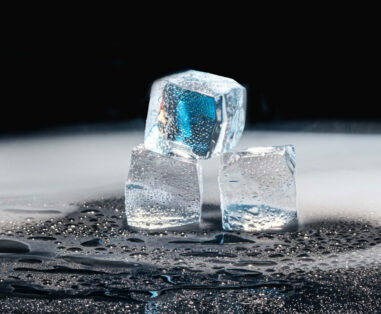 Why Is My AC Freezing Up Frozen Coil Causes Fixes - Tamco Air Three ice cubes stacked on a wet, reflective surface against a dark background, with droplets on the cubes and puddles around them.