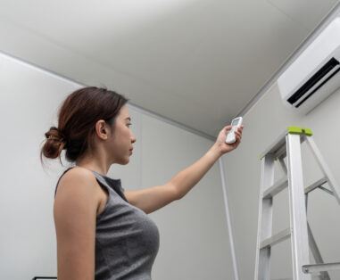 Woman standing beside a ladder, holding a remote toward a wall-mounted air conditioner unit in a bright room.