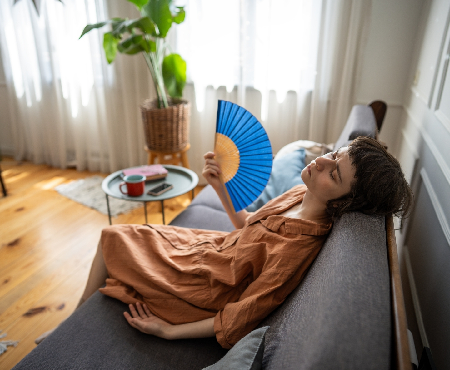 Relaxed woman lounging on a gray sofa in a sunlit living room, fanning herself with a bright blue handheld fan.
